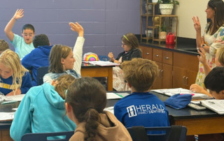 Teacher stands at the front of a classroom calling on students, while several raise their hands during a lesson.
