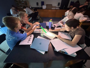 Four students sit around a desk using a flashlight and string for a science activity, writing notes in their notebooks.