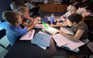 Four students sit around a desk using a flashlight and string for a science activity, writing notes in their notebooks.