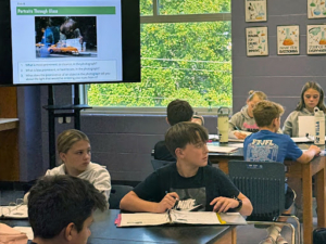 Middle school students sit at tables with notebooks open, looking toward a screen showing a lesson titled “Portraits Through Glass.”