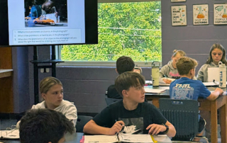 Middle school students sit at tables with notebooks open, looking toward a screen showing a lesson titled “Portraits Through Glass.”
