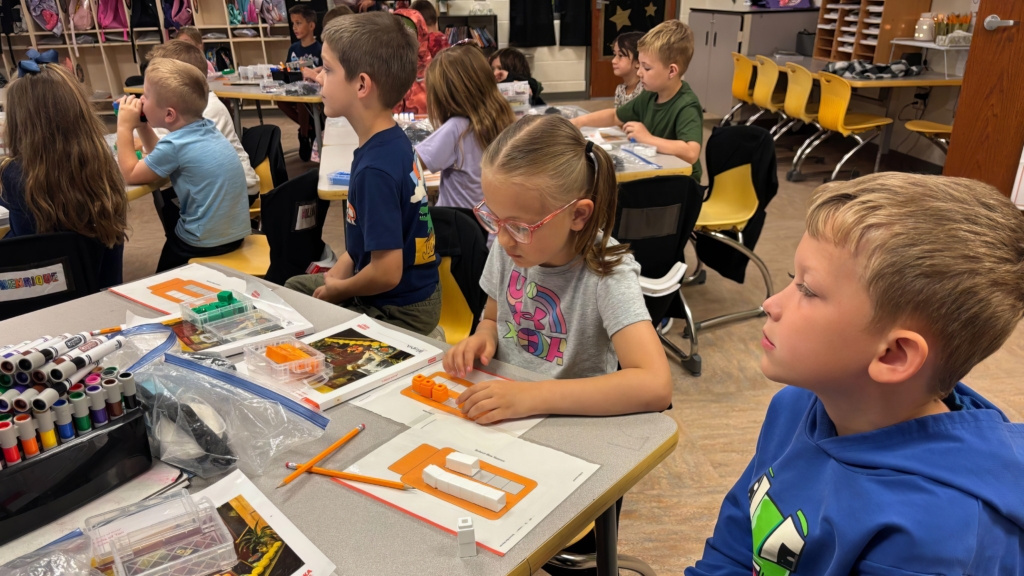 Elementary students focused on a hands-on learning activity at yellow desks in a classroom. In the foreground, a girl with glasses works with small orange and white building blocks while a boy in a blue hoodie looks to the side.