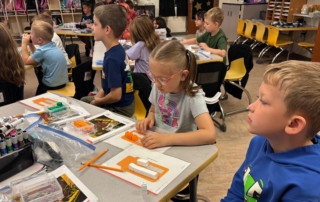 Elementary students focused on a hands-on learning activity at yellow desks in a classroom. In the foreground, a girl with glasses works with small orange and white building blocks while a boy in a blue hoodie looks to the side.