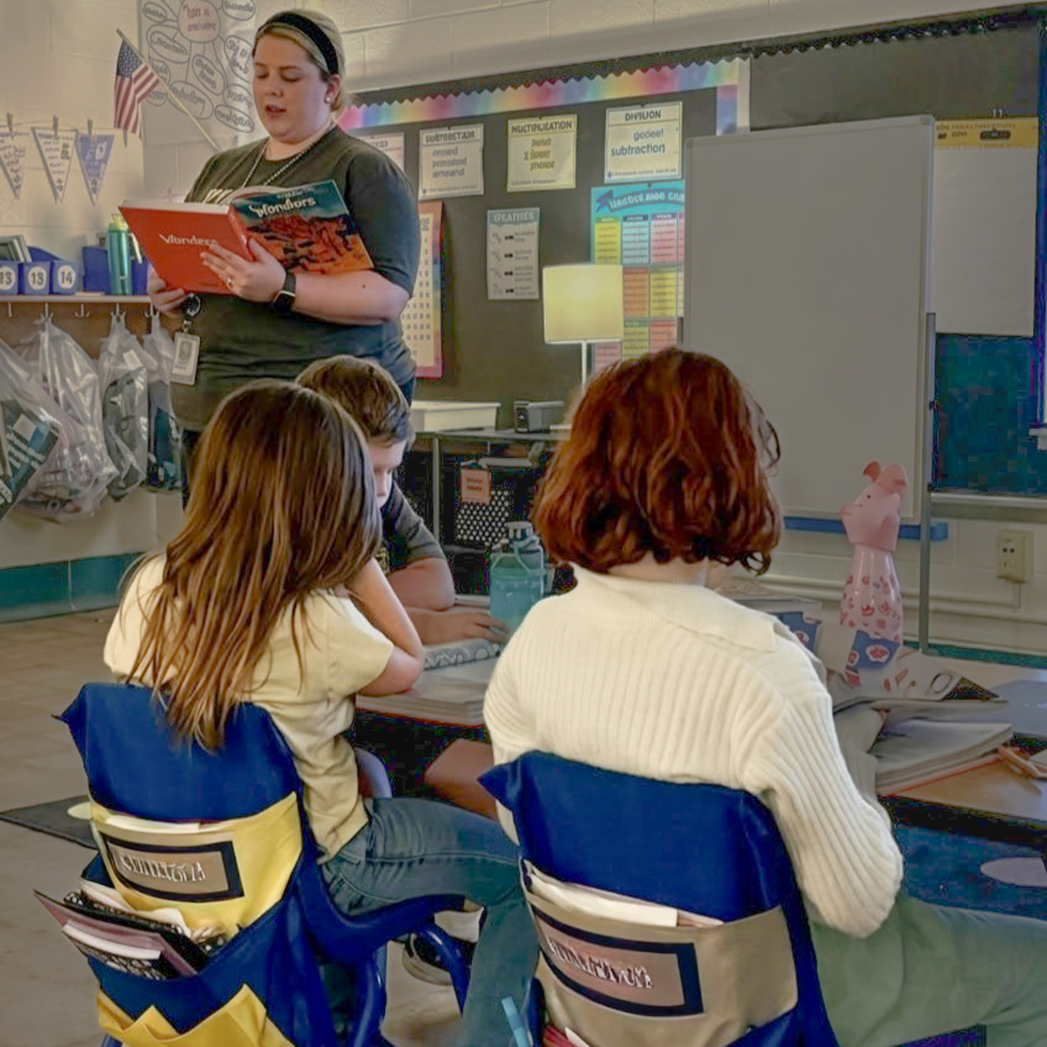 A teacher stands in a detailed elementary classroom, reading from a book titled "Wonders of the World" to three engaged students. The students are seated at blue and yellow chairs with legible name tags, surrounded by clear math and weather educational posters.