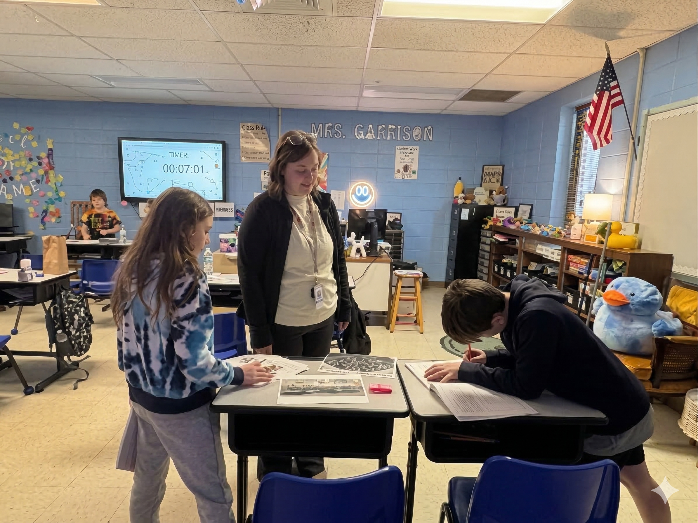 In a blue-walled classroom labeled "Mrs. Garrison," a teacher stands between two third-grade students at their desks. The students are actively engaged in a "Gallery Walk" activity, examining printed historical artifacts and papers. In the background, a digital timer on a large monitor shows seven minutes remaining, and an American flag hangs on the wall.