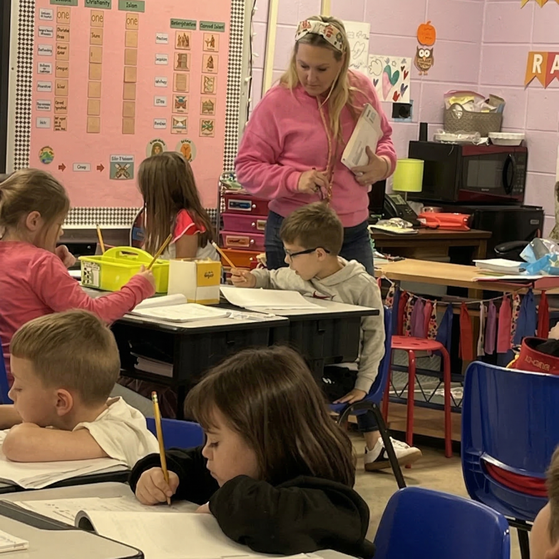 A wide-angle photograph of an active elementary school classroom with corrected, naturalistic colors and balanced lighting. A female teacher with a pattern headscarf, wearing a bright pink sweatshirt, stands centrally, observing a young student in a grey hoodie who is working at his desk. Several students are seated at blue and black desks; some use pencils, others rest on their arms.