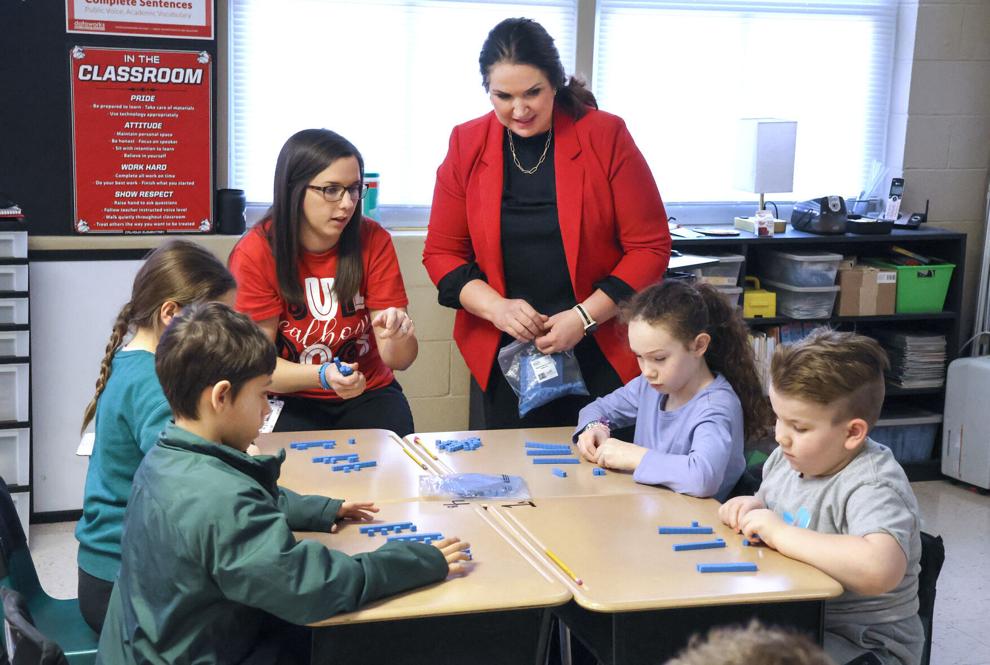 Elementary students work with blue base‑ten blocks at a table while two adults assist, in a classroom with posters, shelves, and natural light.