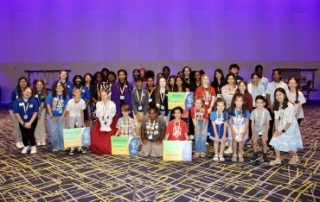 A group photograph of approximately 45 diverse students of various ages posing together in front of a purple illuminated geometric wall and a pattern convention floor. Many wear white JCPS lanyards and some are holding small green or blue signs. A few specific students, including one in a historic costume and others with matching spirit shirts, are visible. The students are smiling and looking at the camera.