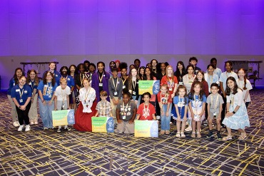 A group photograph of approximately 45 diverse students of various ages posing together in front of a purple illuminated geometric wall and a pattern convention floor. Many wear white JCPS lanyards and some are holding small green or blue signs. A few specific students, including one in a historic costume and others with matching spirit shirts, are visible. The students are smiling and looking at the camera.