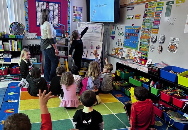 A female teacher stands patiently as a young student points to a sentence on a large wall-mounted monitor during a class on a multi-colored number grid rug. Approximately 15 young children are seated, listening, with one child in the foreground raising their hand. The classroom is filled with educational posters, charts, currency and math displays, and shelving with books and colorful bins. The sentence on the screen reads 'Ron rips paper.'